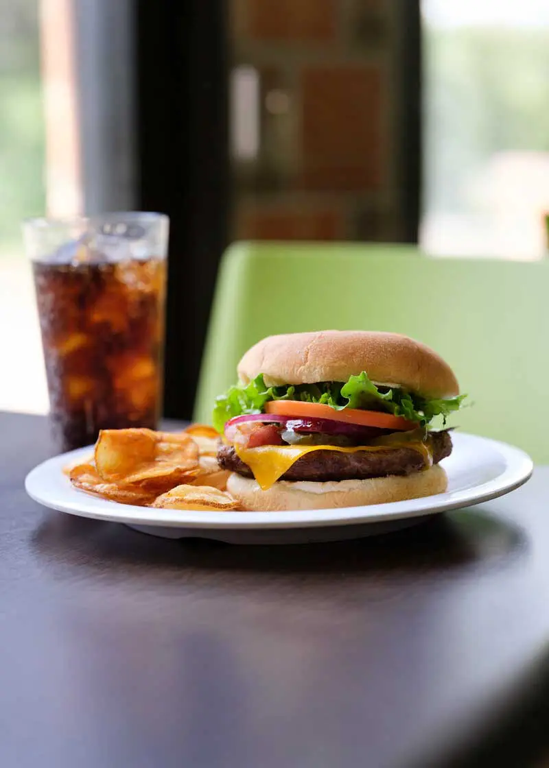 Cheeseburger and chips on a white plate with soda and green chair in the background